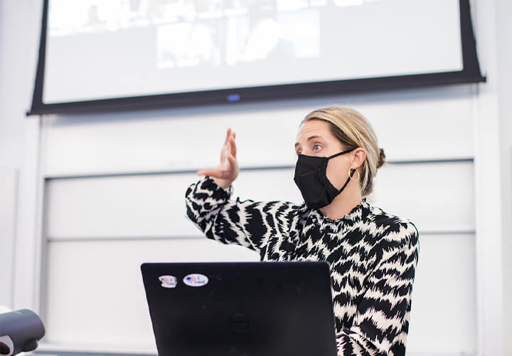 Masked instructor behind a laptop gestures while speaking