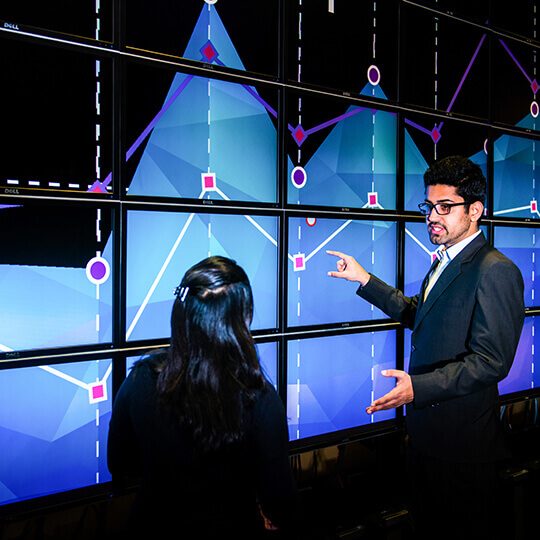 Two people analyze graphs on wall of screens