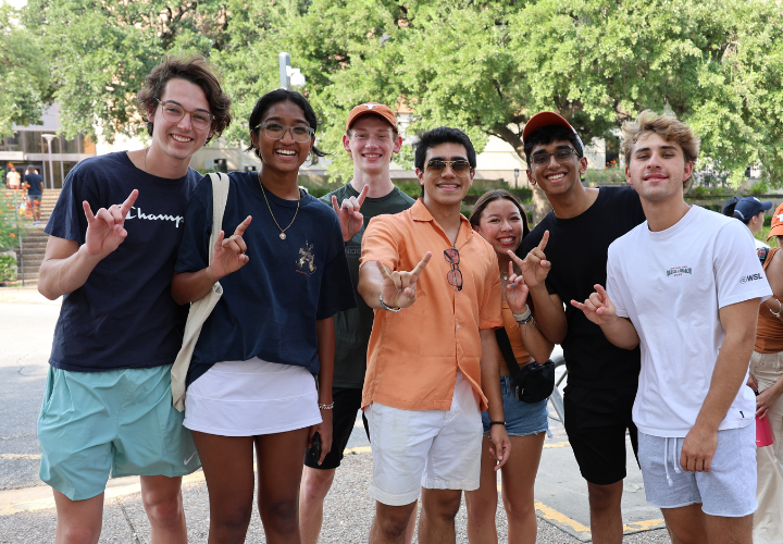 A diverse group of students stand outside smiling and doing hook 'em horns.