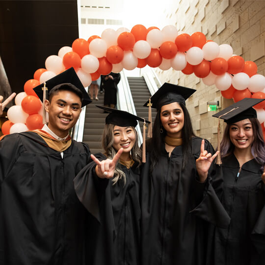 Four graduates in caps and gowns under balloon arch