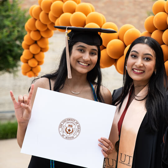 Two graduates pose with diploma in front of balloons