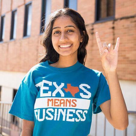 Student wearing a blue UT McCombs t-shirt smiling and giving Hook 'Em Horns